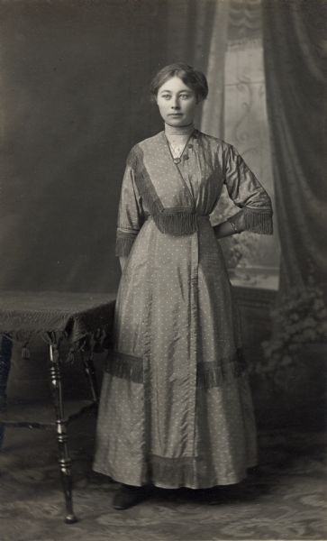 Studio portrait in front of a painted backdrop of a woman standing near a table wearing a dress with her hand held behind her back. The dress has fringe detailing.