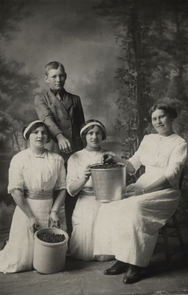 Studio portrait in front of a painted backdrop of four young people. From left to right: Teresa Kalscheur Endres, Frank Kalscheur, Julia Kalscheur, and Teresa Kalscheur, posing with a crock and a bucket of blackberries. The two Teresas are cousins. The two women on the left are wearing ribbons in their hair.