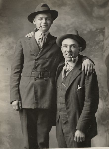 Studio portrait in front of a painted backdrop of two men, Edward Kerd on the left and Matt Valentine on the right. Edward appears to be standing on a stool. Each man is wearing a suit, tie, and hat, and has a pipe in his mouth.