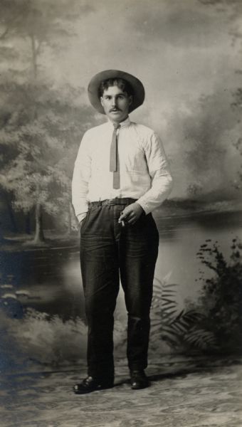 Dressed in a shirt, hat and narrow tie, Charles Brendier poses for a studio portrait in front of a painted backdrop with a cigar in his hand.