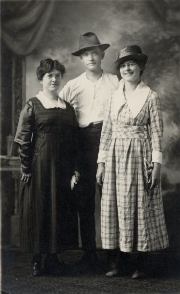 Studio portrait in front of a painted backdrop of two women standing on either side of a man wearing a hat. Both woman are wearing dresses, and the woman on the right is also wearing a hat.