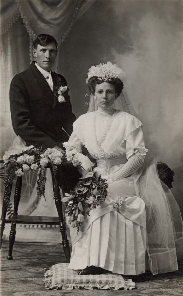 Studio portrait in front of a painted backdrop of a bride and groom on the occasion of their wedding. The groom, Matt Koch (son of Henry Koch), is sitting on a table behind his bride, Christina Bergman Koch, who wearing a white wedding dress, and is sitting in a chair and resting her foot on a pillow.