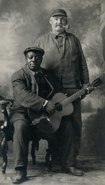 Two men posing for a studio portrait in front of a painted backdrop. James Gorman, standing, has a pipe in his mouth; the seated man holding a guitar is unidentified.