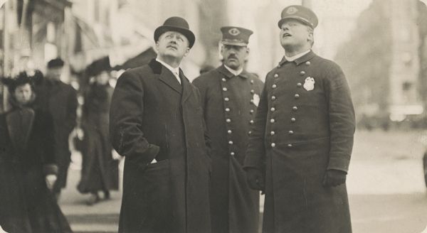 A businessman, dressed in a suit coat and hat looks up while standing near two police officers. Behind them is a crowd of pedestrians on a street.