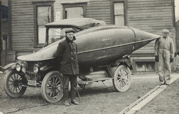 A man poses in front of a canoe tied onto his car. A second man looks on from the background.