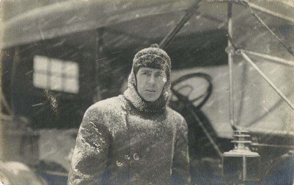 Winter scene with a man dressed in a winter coat and hat standing in the snow in front of a car.