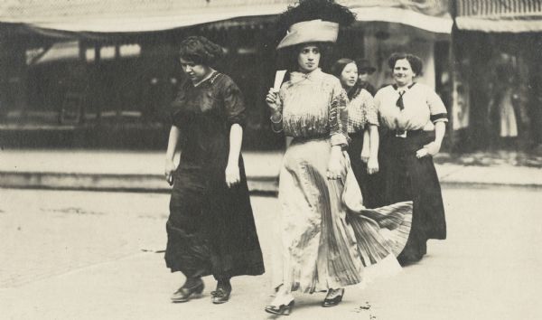 Four women crossing a street. One of the women in the foreground is dressed in a fashionable dress and hat.
