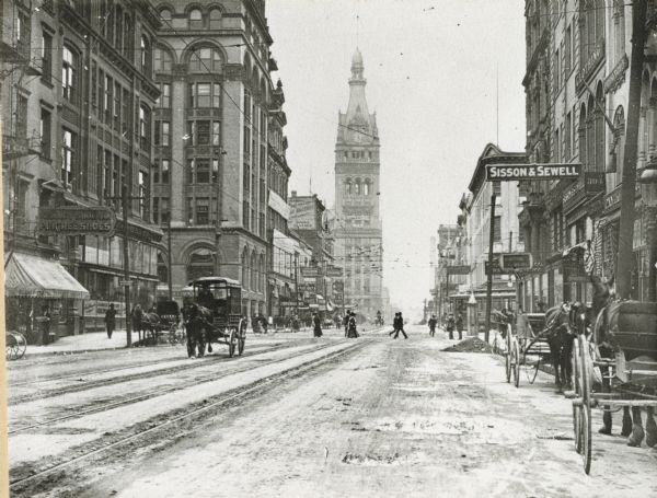 North Water Street with City Hall in the background. Numerous horse-drawn vehicles are driving along the street and parked along the curbs, and pedestrians are crossing the street.