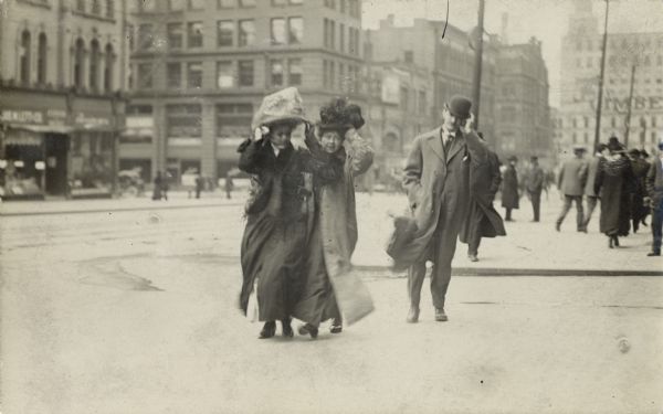 Two women and a man in the foreground hold onto their hats on a windy day as they cross a street.