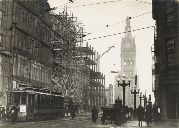East Water Street at Wisconsin Avenue. City Hall is in the background. At the left is the Waldheim and Company Department Store of 407-411 East Water Street, and Swoboda's Cafe at 432 East Water Street. Buildings are under construction further down the street on the left. At the right are the Law Offices of 82 W. Wisconsin Avenue.