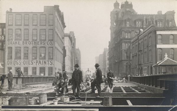 Men at work on a bridge under construction. A sign on a building in the background reads: "Drake Brothers Co." and "Drugs • Oils • Paints."