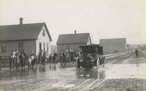 An automobile is attempting to cross a flooded street as onlookers are watching.