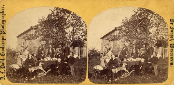 Reverend Carl Christian Aas (1843-1898) and family gathered outside around a table. There is a rocking horse and baby carriage in the view. The stone house was the first parsonage for the Lutheran Church at Wiota.