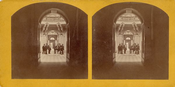 Stereograph of view from a first floor hallway into the rotunda of the third Wisconsin State Capitol where a group of men are standing. Published specifications for the 1882 addition tells us that the main hallways resembled this floor: "The halls, vestibules, and corridors of the principal floors to be tiles with white marble and blue slate tiling twelve inches square and one inch thick, with a slate border to correspond to the old floors... The floors of the connecting corridors of the second and third floors to be floored with Minton tile, six-inch octagon, with two inch corner tiling of colors to correspond to the old floors."
