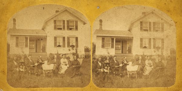 Seven individuals sit around a table on a front lawn. The four men on the left of the table sit with open books.