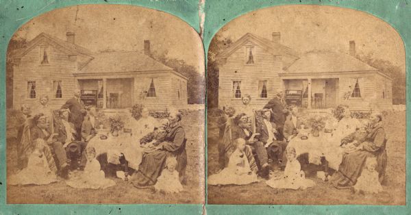 A family is posed around a table on which are many thick books and a stereoscope. Most of the men, especially the twin boys, have assumed heroic postures. The frame house in the background has a piano and a sewing machine on its porch. Farm buildings are in the far background.