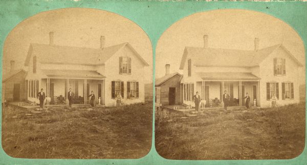 Two men, two women are posing in front of a frame house that has a chair, sofa, table and a small white statue on its front porch. There are three doors and one window on the porch. Cut work is above the windows on the left side and right front of the house. On the left is a birdhouse and a dog on an open platform walkway on the foundation that leads to another building at the corner of the house, which is built into a hill. Stereograph print has text on back that reads: "John Skake(?) family, Wiota".