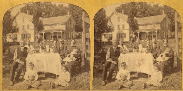 A family of nine sits on their front lawn with the interior tableware set for tea or coffee. In the background, a man is leaning against an unidentified object.
