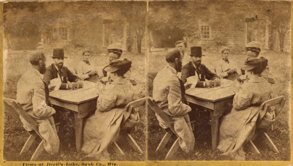Stereograph of a group of men and women sitting outdoors, playing a card game around a table at Devil's Lake.