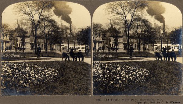 View of the Fourth Ward Park. Several men sit on a bench, while numerous houses and a factory are visible in the background.
