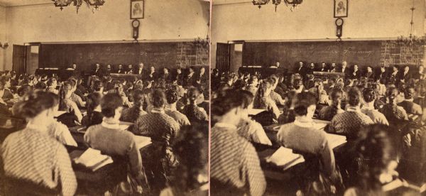 A full room of students at the State Normal(?) School. All of the students appear to be female. A line of possible teachers are in front of the blackboard.