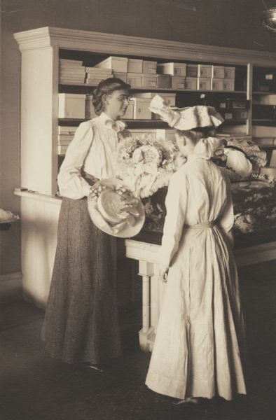Fontana A. Massee, member of the Menomonie High School class of 1905, depicted as a milliner. Pictured showing hats to another girl. Part of a yearbook created by classmate Albert Hansen, based on a class prophecy theme.