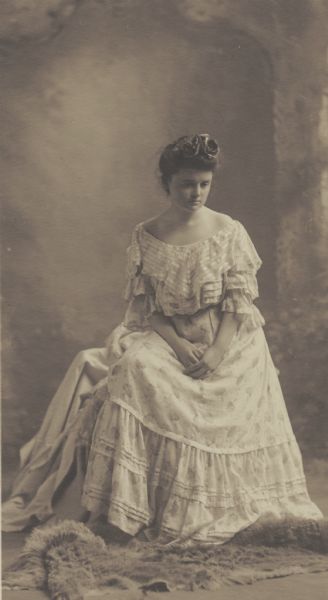 Susan M. McCutcheon, member of the Menomonie High School class of 1905, depicted as an actress, posing on a fur rug. Part of a yearbook created by classmate Albert Hansen, based on a class prophecy theme.
