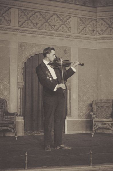W. Lambert Clark, member of the Menomonie High School class of 1905, depicted as a violinist. Pictured in a tuxedo, playing a violin. Part of a yearbook created by classmate Albert Hansen, based on a class prophecy theme.