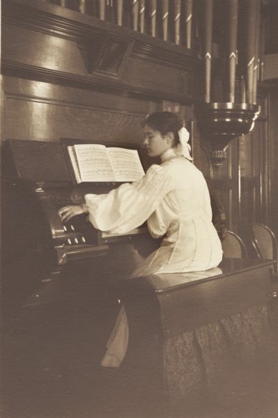 Katherine M. Harmon, member of the Menomonie High School class of 1905, depicted as a pianist, playing an organ. Part of a yearbook created by classmate Albert Hansen, based on a class prophecy theme.