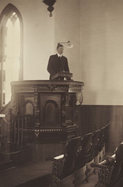 Paul Toft, member of the Menomonie High School class of 1905, depicted as a preacher, at a pulpit. Part of a yearbook created by classmate Albert Hansen, based on a class prophecy theme.