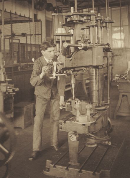 Robert E. Clough, member of the Menomonie High School class of 1905, depicted as an inventor, using a large drill press. Part of a yearbook created by classmate Albert Hansen, based on a class prophecy theme.