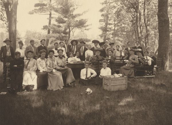 Group portrait of the senior class at Menomonie High School from 1905, having a group breakfast at Riverside Park. A crate with food on it appears in the foreground. Part of a yearbook created by classmate Albert Hansen, based on a class prophecy theme.