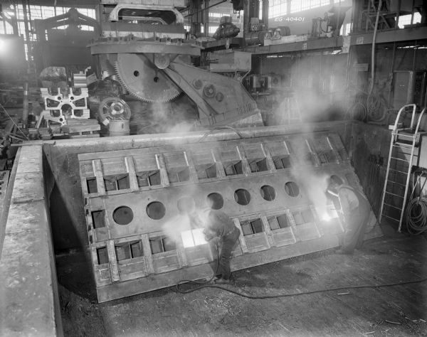 Two men simultaneously weld. According to an original Falk caption, "rough weight. 72000 pound est.-Unit was rough machine to with <i>[sic]</i>. 1/4 inch of finish size & second stress relieved by Falk. This with mating <i>[sic]</i> smaller runway is largest horizontal boring mill in U.S.A."