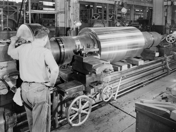 A man oversees the manufacturing process of a roll drive pinion which was purchased by E.W. Bliss Company of Salem, Ohio, and used by Empire Steel Company. Falk original caption reads: "Turning of Journal Diameter. 38/28 teeth, 1 DP, 72 inch face, 3-3/8 inch gap, 23 degree full depth tooth form, staggered herringbone teeth, 23 degree helix angle, 4.63 axial overlap, 38.000 inch pitch diameter, 39,040 inch outside diameter, 24 inch shaft diameter, 180 inch shaft length. Pinions were manufactured from AISI 4350 forging heat treated to 305-355 BHN. Rough weight of pinion forging was 40,000 pounds each. Peak load transmitted by pinions is 6250 HP from 50 to 50 rpm. Pitch line velocity is 498 ft. per minute. AGMA surface durability rating is 6220 HP. Pinion drawing numbers are 318352 and 318353. Price per set of two pinions is $56,250. Pinions were ordered on Bliss purchase order SA 75165 dated December 18, 1956. Pinions were quoted on Falk inquiry E5693-0790B."