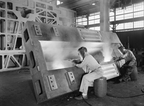 Two seated men weld a sugar mill drive that was later purchased by E.L. Dennis Engineering Company of New Orleans, Louisiana and used by A. Wilbert's Sons Myrtle Grove Factory of Plaquemine, Louisiana. Original Falk caption reads: "Welding of oil sump to base. Base drawing 411353. Unit Size: 75.833 inches x 24 inches E.L. Dennis Engineering Company purchase order number 13991 dated November 27, 1956."