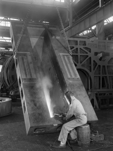 A seated man welds a sugar mill drive that was later purchased by E.L. Dennis Engineering Company of New Orleans, Louisiana and used by A. Wilbert's Sons Myrtle Grove Factory of Plaquemine, Louisiana. Original Falk caption reads: "Welding of oil sump to base. Base drawing 411353. Unit Size: 75.833 inches x 24 inches. E.L. Dennis Engineering Company purchase order number 13991 dated November 27, 1956."