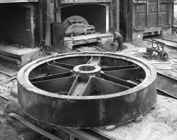 A large Drive Gear rests outside the factory after being removed from the furnace. This product was later purchased by Buffalo Forge Company of Buffalo, New York, and used by Sociedad Agricola Pucala Ltda, in South America of Lima, Peru. Original Falk caption reads, "Gear Casting being removed from Annealing furnace. 26-158 teeth, 1 DP, 24 inch face, 3 3/8 inch gap, 23 degree full depth tooth form, staggered herringbone teeth, 23 degree helix angle, 1.39 axial overlap, 92.750 inch center distance, 6.0769 to 1 ratio. The pinion and gear set is normally required to transmit a load of 650 HP from 36.69 to 5.99 rpm. The pitch line velocity is 249.7 feet per minute."