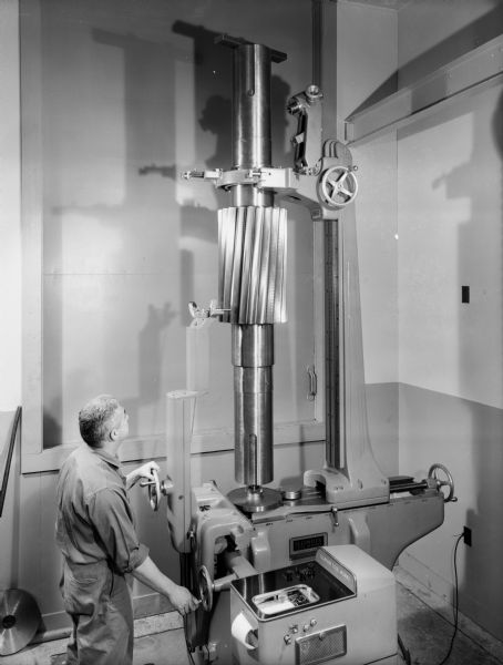 A male employee operates a large machine at the Falk Corporation. The gear was purchased and used by the Falk Corporation. Original Falk caption reads: "The machine and recorder are manufactured by Illinois Toolworks of Chicago. The machine is capable of checking the accuracy of involute profiles on pinions or gears up to 36 inch outside diameter. Pinion description: 20 teeth, 1 1/2 DP, 24 inch face, 5 degree helix angle, single helical, full depth mill type teeth, right hand helix. The pinion is shown on drawings 231189 and 411363."