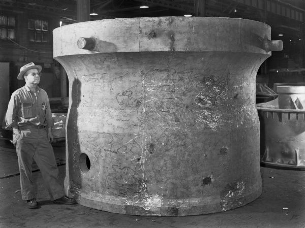 A male employee stands next to a large shipping shield. This shield was purchased by Stearns-Rogers and used by Westinghouse. Original Falk caption reads, "Upper half casting-55,700 pounds-rough dimensions. 103 inch OD and 99 1/2 inch OD x 95 inch ID and 87 inch ID x 81 inches long."