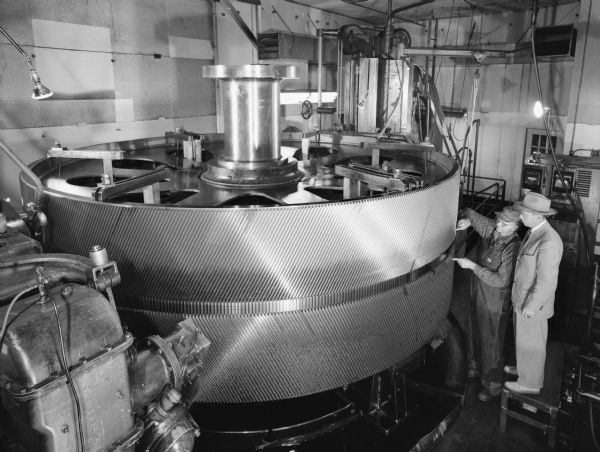 Two men inspect a large gear which was later purchased by DeLaval Steam Turbine Company of Trenton, New Jersey. Original Falk caption reads, "This gear is shown on Falk Drawing 319935. The gear blank and hobs were furnished by DeLaval. The gear has 610 DeLaval HB teeth of 3.38779 DP, 55 1/2 inch face, 3 1/2 gap, 27 degree helix angle, 180.059 pitch diameter equals 3.811687 and the normal circular pitch equals .824202. Note the 55 1/2 face width."