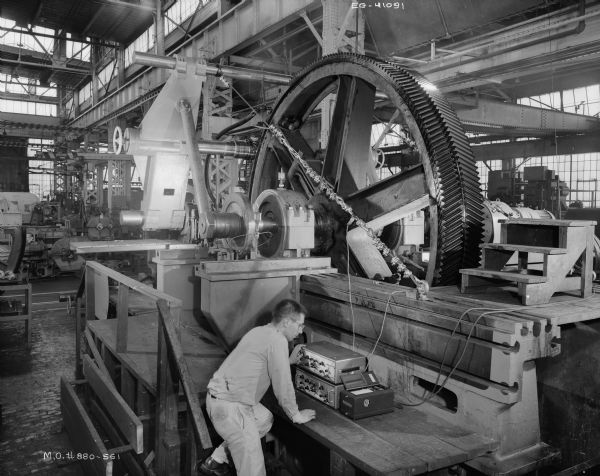 A man attends to a control panel of a factory machine that tests a large gear. The Falk Corporation later purchased and used this gear. Original Falk caption reads, "Gear: 229 teeth, 1 1/2 2DP, 23 degree full depth herringbone, 20 inch face width, 152,667 inch pitch diameter, 153.893 outside diameter, 17.869 inch bore, medium carbon cast steel annealed, rough weight is 26,260 pounds. Split rim and hub, drawing 407932, pattern 11485 H A/B. The gear was originally sold to Nordberg Manufacturing Company on M.O. 460-119 in 1951, and replaced in 1955 by a gear manufactured on M.O. 5545-0065. The integral with shaft pinion is similar to the pinion shown on drawing 317102, and was salvaged after manufacturing errors made it unsuitable for the original order. Pinion P.D. is 14.133 inch, O.D. is 15.359 inch, 21 teeth."