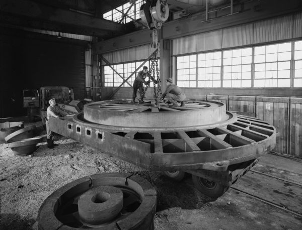 Two men crouch atop of the large gear, while another stands at the side overseeing the lift. This gear was purchased and used by the Ladish Company of Cudahay, Wisconsin. Original Falk caption reads, "Weight: 120,060 pounds (center section), 24 feet in diameter and 17 feet across flats."