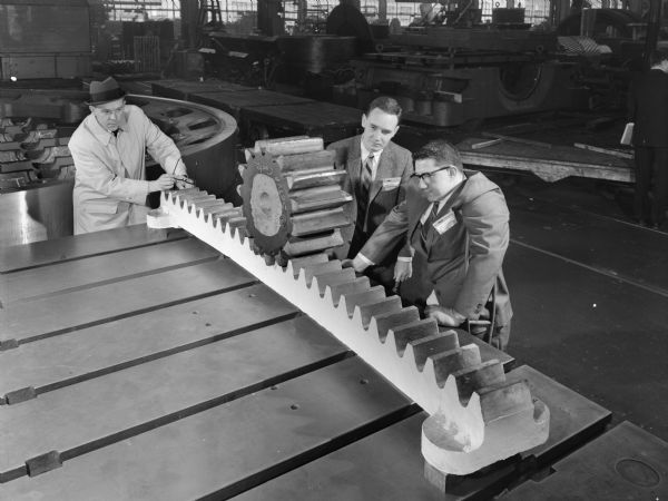 Three men with bevel gear and pinion that was later used in a Rotary Hearth Furnace drive. This gear was purchased by Surface Comb. Division of Midland Ross and used by the National Steel Company. Falk caption reads, "Segment approximately 8 feet long, 7 inches high with 26 bevel cast teeth having a 7 inch face and 3.8 circumference pitch. Pinion approximately 21 1/2 O.D. and 15 cut bevel teeth with 9 inch face. 30 segments make one 80 foot diameter. Bevel gear ring in mesh with 3 pinions transmitting 25 HP, each at 90 feet per minute. Total of 780 teeth in gear, 3.8 inch circumference pitch x 7 inch face. 15 cast teeth x 9 inch face in pinion. The rotary furnace, 100 feet O.D. and 60 feet I.D. is driven by 3 2090 Y3 units through footing shaft, "P" type couplings connected to the bevel pinions." 
   The men in the photograph are Frank Rinker, Chief Engineer of Minerals Processing, and Jim Gill and Art Keel, both Project Engineers.
