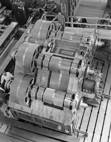 View from above of double reduction-reverse gear that is a component of a gas turbine drive. This gear was purchased by Sun Shipbuilding and Dry Dock Company. Falk caption reads, "Partially assembled unit showing gear train and general arrangement." Male employees in photograph are L. Goreson and D.J. Feilbach.
