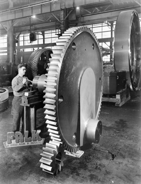 Tire curing press that was later purchased by McNeil Akron Corporation. Falk caption reads, "14-90T, 10 P Spur, 7 inch face. Crank gears have two unique features, not previously preformed at Falk. 1. Stress distribution groove. 2. Cold roll finish on bearing diameter and radii." Male employee in photograph is J. Martorena.