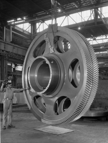 Bull gear for shaft mounted on a BOF drive. This gear was later purchased by Pennsylvania Engineering Corporation and used by Youngstown Sheet and Tube. Falk caption reads, "These photos taken for use as photo/caption release material prior to 1968 Steel Show. This gear, with four pinions fastened to it, constitute the predominant element in the Falk exhibit at the show." Male employee in the photograph is George Dickinson.