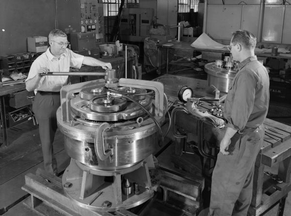 Two Falk employees, Walt Drobkia on the right and Norb Winski on the left, attend to a planetary gear drive that is a component of a centrifuge. This product was purchased by Combustion Engineering Incorporated and used by Black Mesa Pipe Line. Falk caption reads, "These photos illustrate various phases of assembly and testing of a special planetary cone centrifuge and identified as their number 4080 dyna cone."