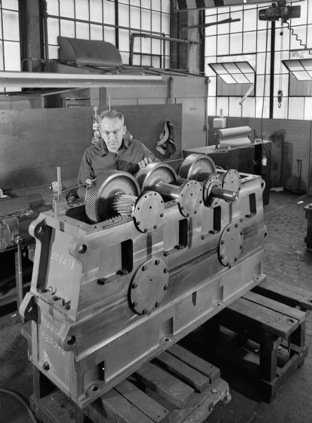 A male employee, Chet Nycz, attends to an output shaft that was purchased by the Road Machine Division of American Hoist and Derrick Company and used by Vigus Quarries of Bridgeton, Missouri. Original Falk caption reads, "Special double reduction, dual output speed reducer also furnished with 14 feet couplings and 190F's on LS shafts... see Engineering File for exact engineering details."