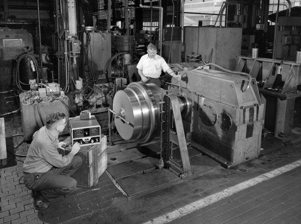 Planetary gear drive that was used as a later component in a centrifuge. This product was purchased by Combustion Engineering Incorporated and used by Black Mesa Pipe Line. Falk caption reads, "Photo of test arrangement in Shop 4 for combustion units... for exact engineering specifications see Engineering File." Male employee on the right is Norb Winski and the male employee on the left is John Grudichak.