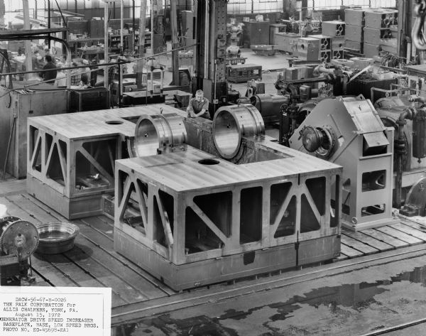 This speed increaser, a component of a generator drive, was purchased by Allis Chalmers and used by the United States Corps of Engineers. Falk caption reads, "Low level dam site/generator drive. Large special speed increaser for Ozark dam site on Arkansas river... see Engineering File for exact details. This view shows unit base with base plate/base/and low speed bearings." Male employee in photograph is Norb Winski.